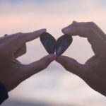 hands holding heart-shaped shell on beach at sunset